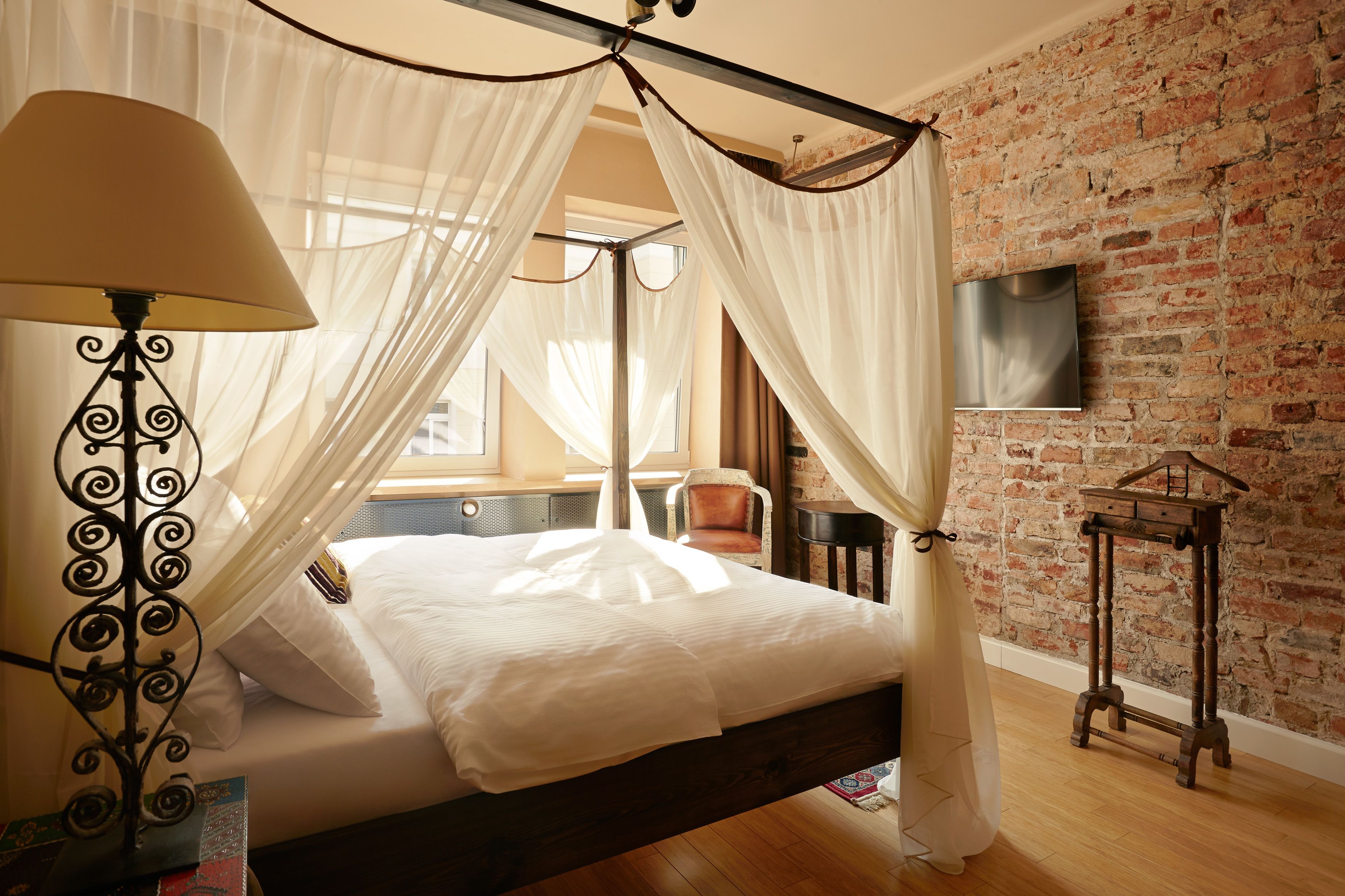Un dormitorio con cama con dosel, una lámpara de noche, pared de ladrillo y televisor en la pared.