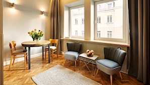 A modern living room with a wooden floor, a round table with chairs, a small side table with fruit, and two windows with curtains. - SleepWell Apartments Ordynacka (Warsaw)
