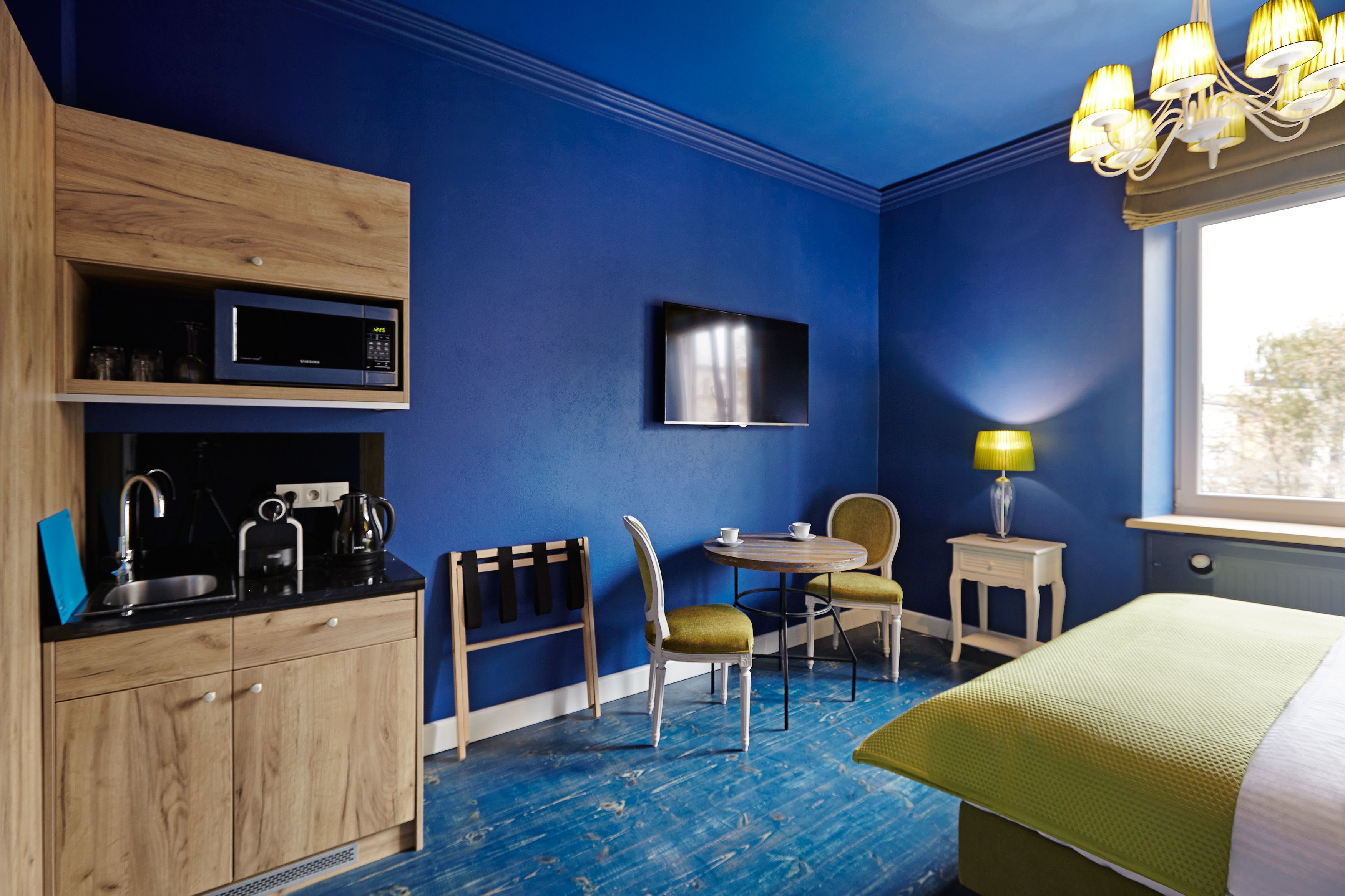 A compact hotel room with a blue wall, a flat-screen TV mounted above a wooden cabinet, a small dining table with two chairs, a bed with a green bedspread, and a window with a view of trees.