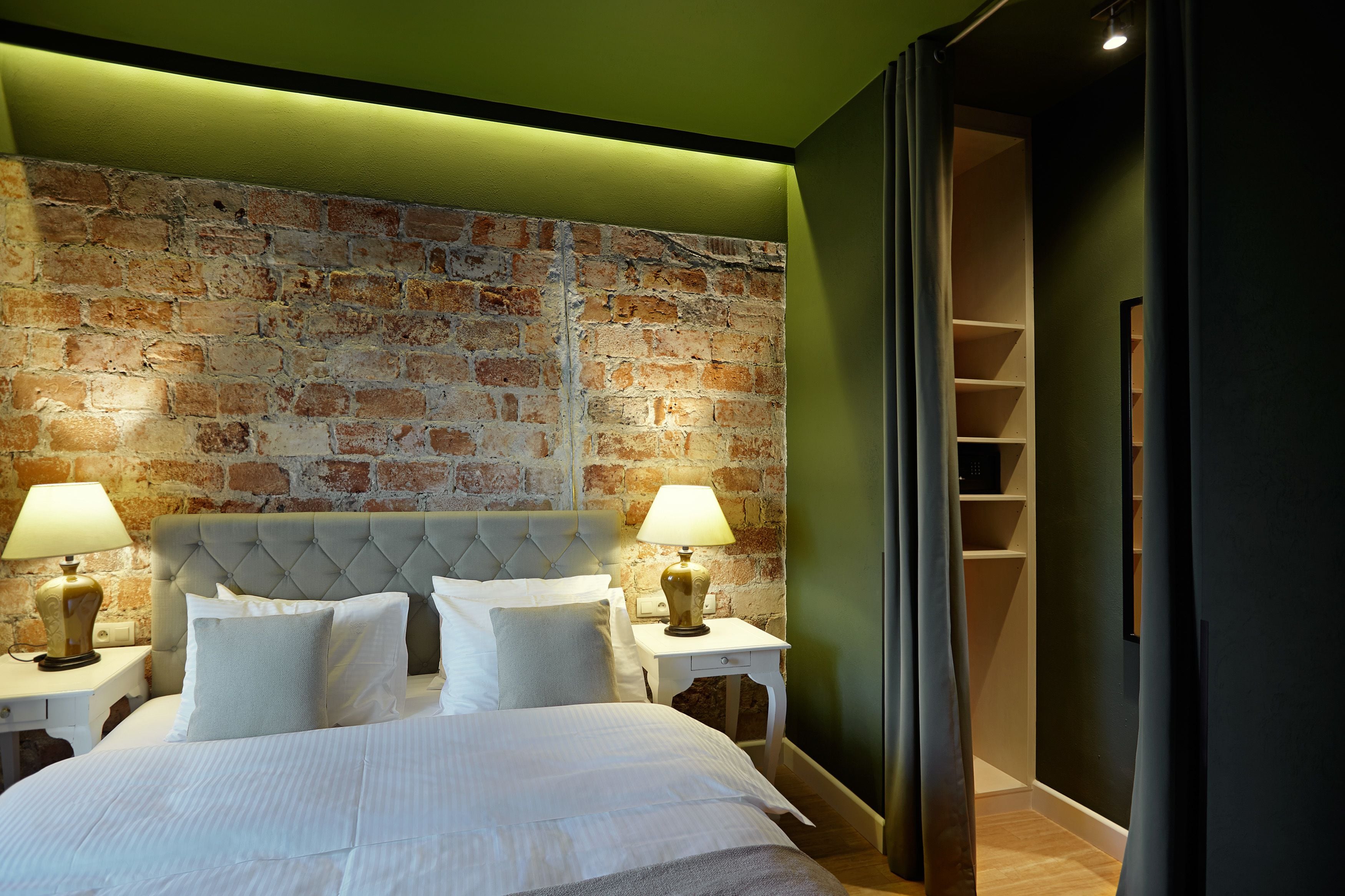 Habitación de hotel con pared de ladrillo, cabecero capitonado, cama con ropa blanca, dos mesitas de noche con lámparas y un armario empotrado con estantes.