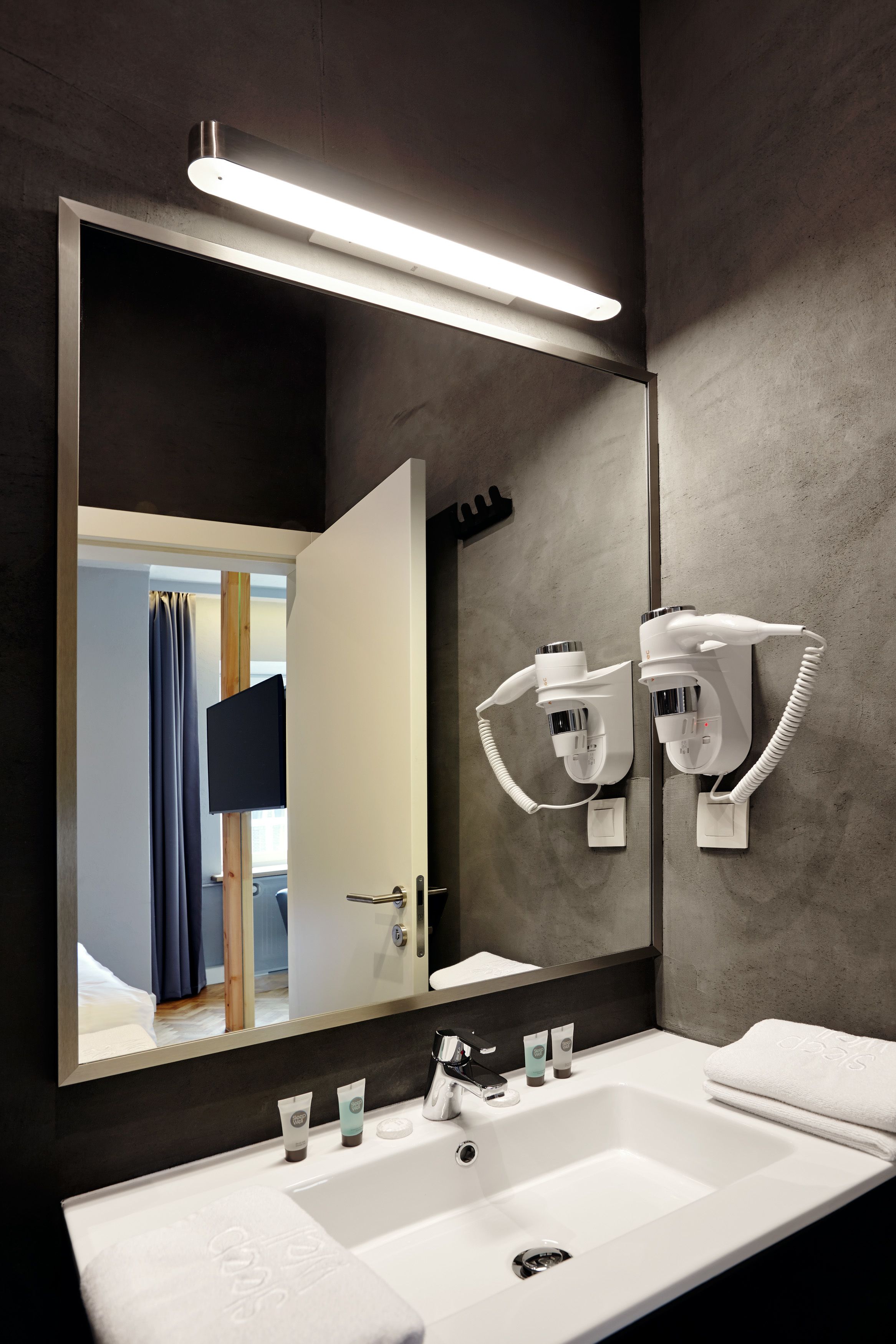 A modern bathroom with a large mirror, a white sink, and two hair dryers mounted on the wall.