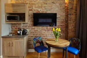 Una cocina compacta con microondas, cafetera y hervidor, un televisor de pantalla plana montado en una pared de ladrillo, y una mesa redonda de madera con dos sillas azules.