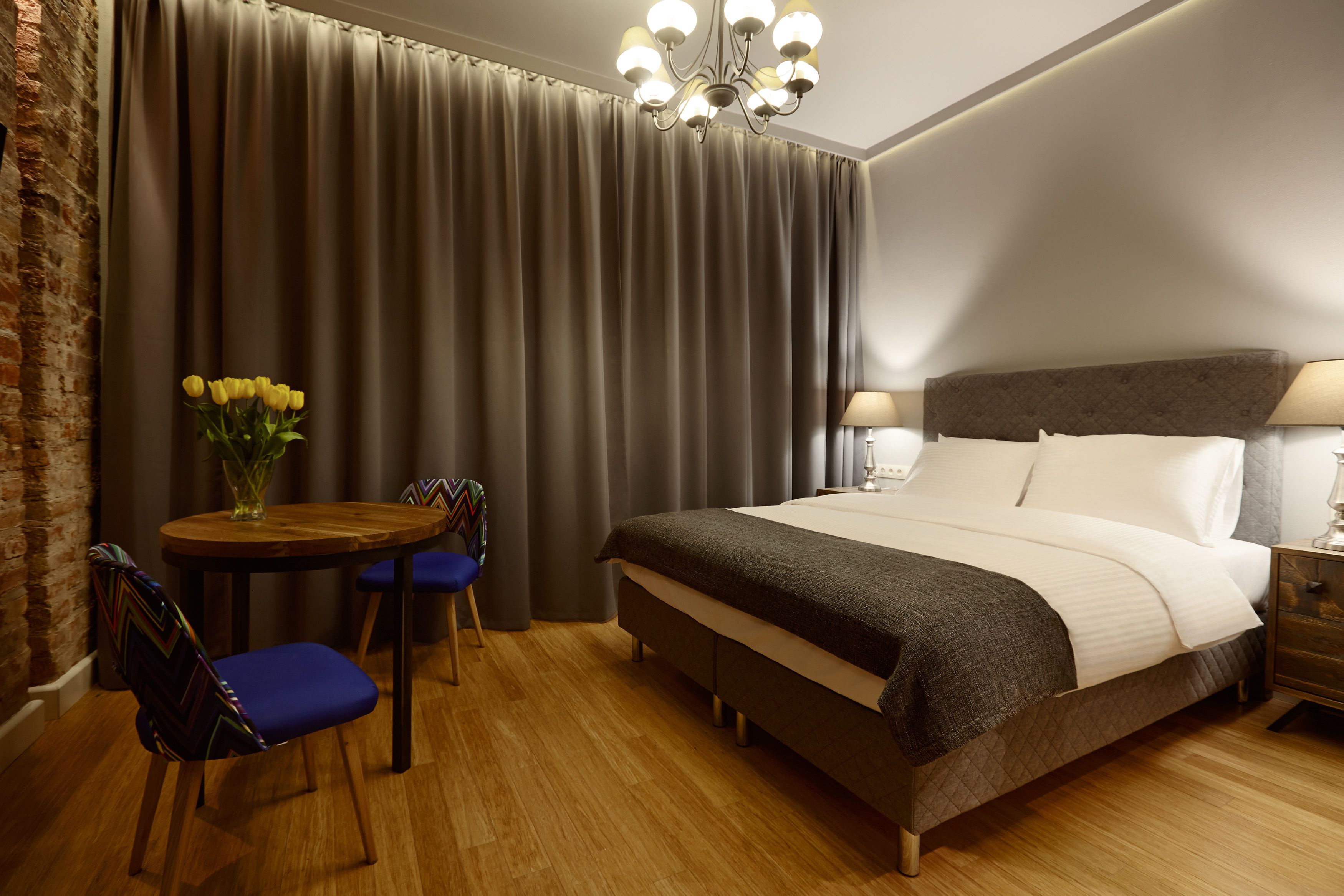 Ett hotellrum med en stor säng, ett träbord med stolar, en vas med gula blommor och en kristallkrona.