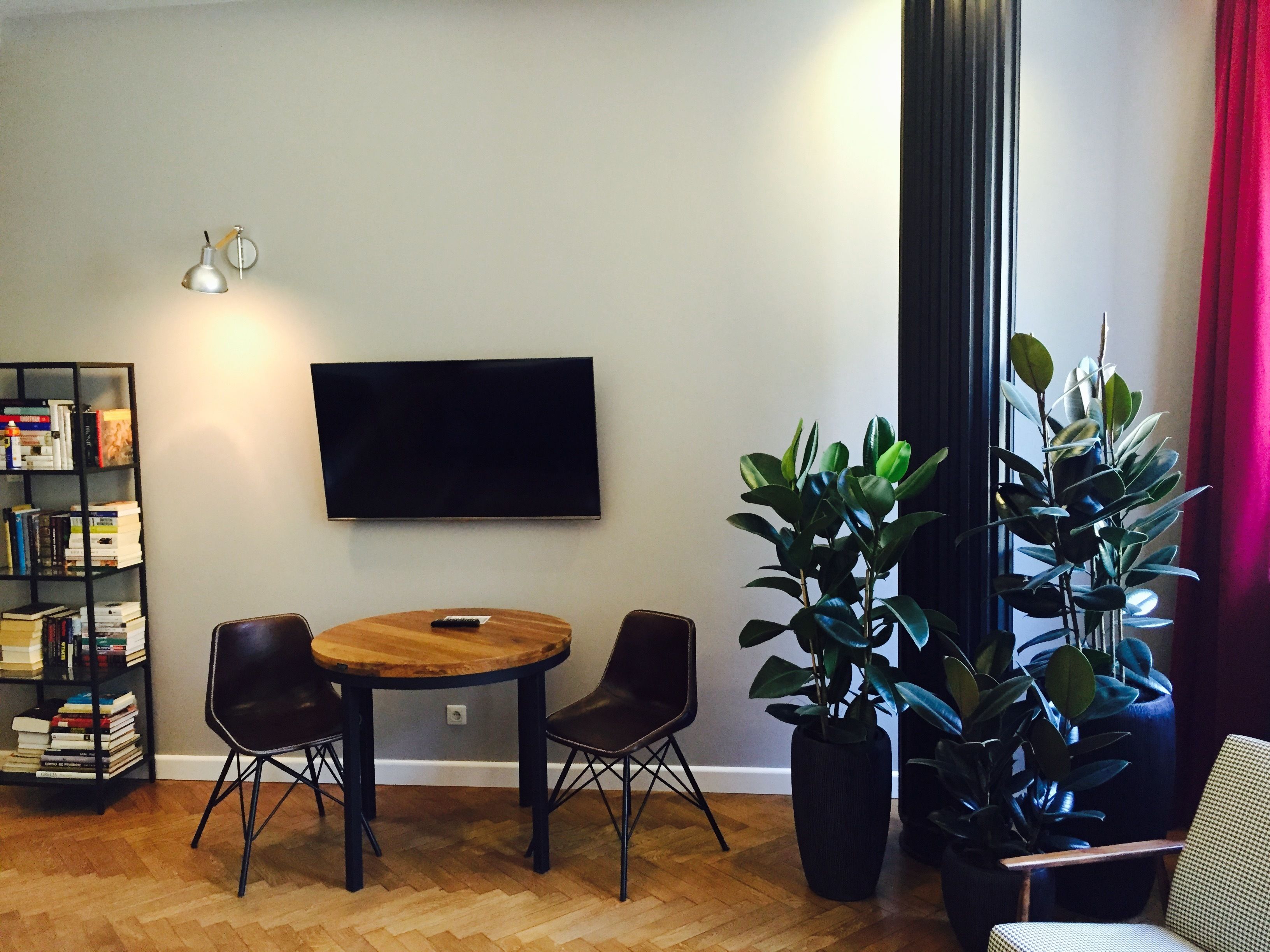 A living room with a round wooden table, two black chairs, a bookshelf, a flat-screen TV, and two large potted plants.