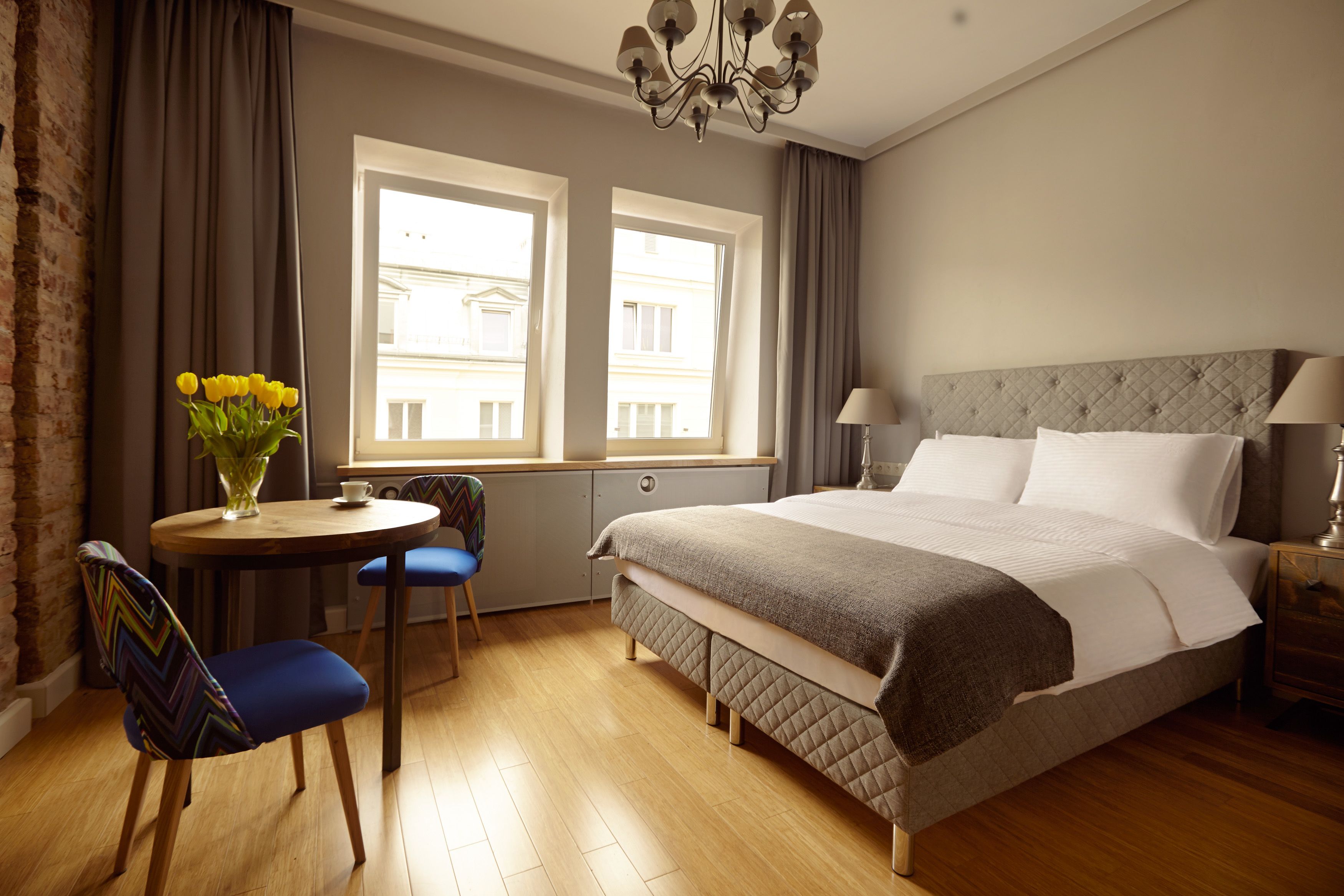 Une chambre d’hôtel avec un lit, un bureau et une chaise, un vase de fleurs et une fenêtre donnant sur des immeubles.