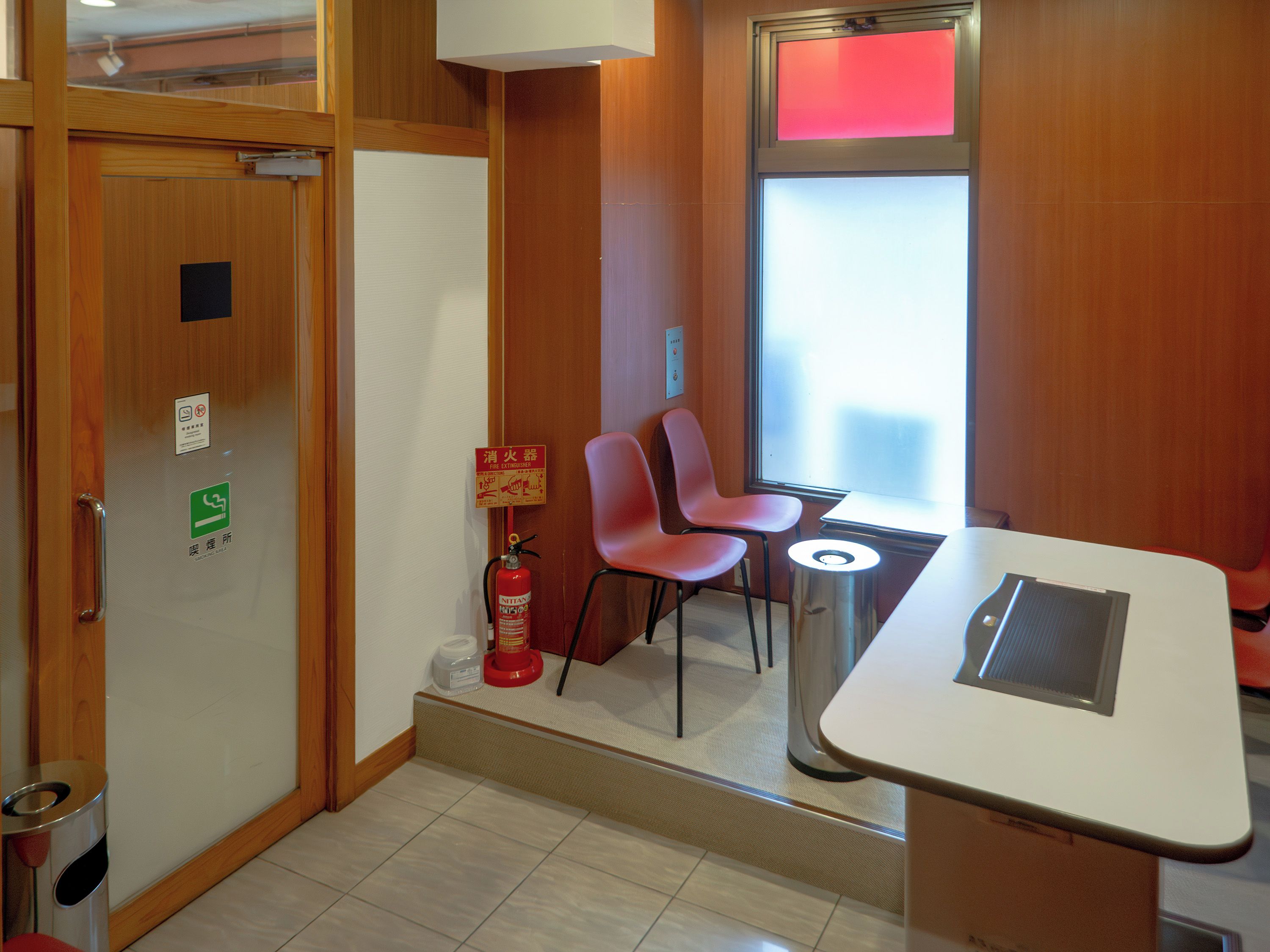 Una camera d'albergo con una porta in legno, un estintore, un tavolino con una sedia e una finestra con una luce rossa.