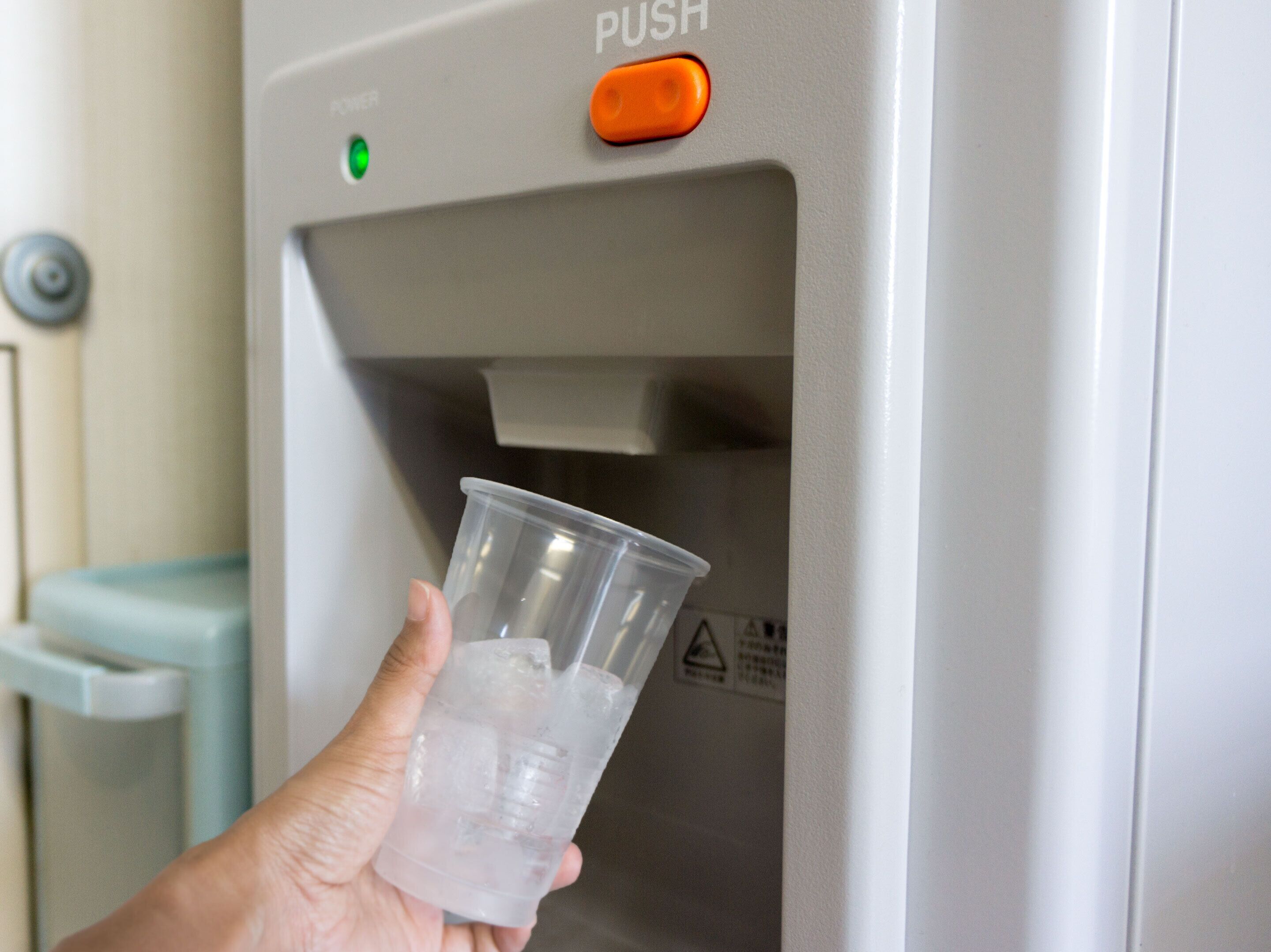 Une main tenant un gobelet en plastique rempli de glaçons, devant un distributeur d’eau doté d’un bouton d’alimentation vert et d’un bouton-poussoir orange.