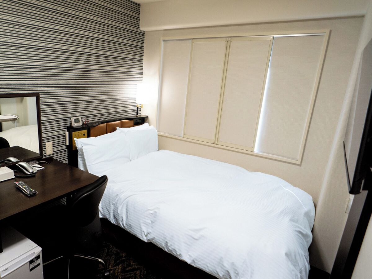 A hotel room with a bed, a desk with a telephone, a mirror, and a window with blinds.