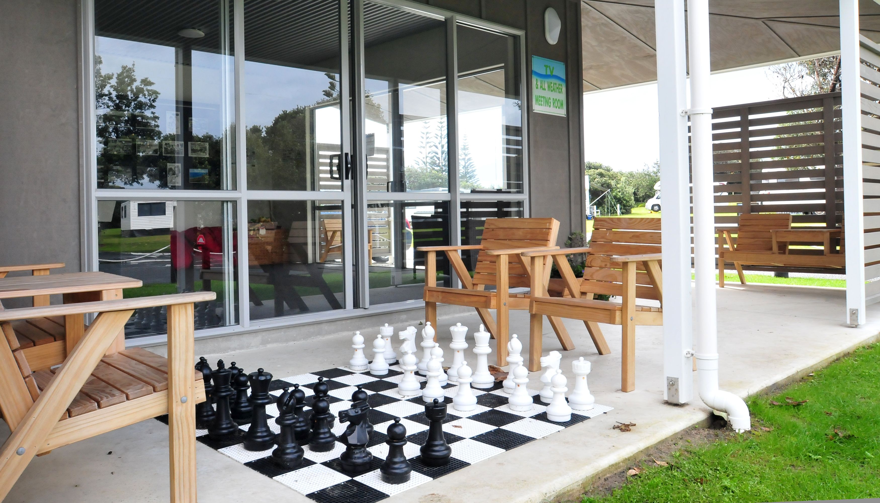 Un jeu d’échecs est installé sur une surface en béton, à l’extérieur d’un bâtiment doté de bancs en bois et d’un panneau indiquant la présence d’une salle de réunion équipée d’un téléviseur.