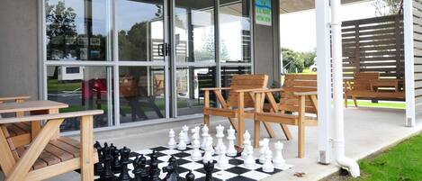 Un jeu d’échecs est installé sur une surface en béton, à l’extérieur d’un bâtiment doté de bancs en bois et d’un panneau indiquant la présence d’une salle de réunion équipée d’un téléviseur.