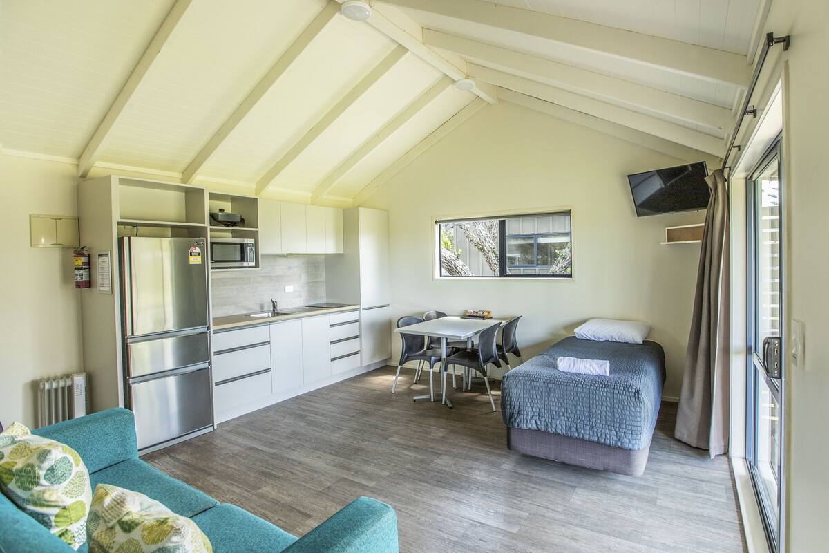 A compact room with a kitchenette, a dining area, a bed, and a TV mounted on the wall.