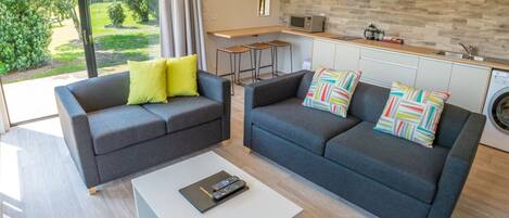 A modern living room with two grey sofas, a white coffee table, and a kitchen area with a stone backsplash and a washing machine.