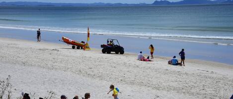 A beach with people sunbathing and playing, a lifeguard tower, and a mountain in the background.