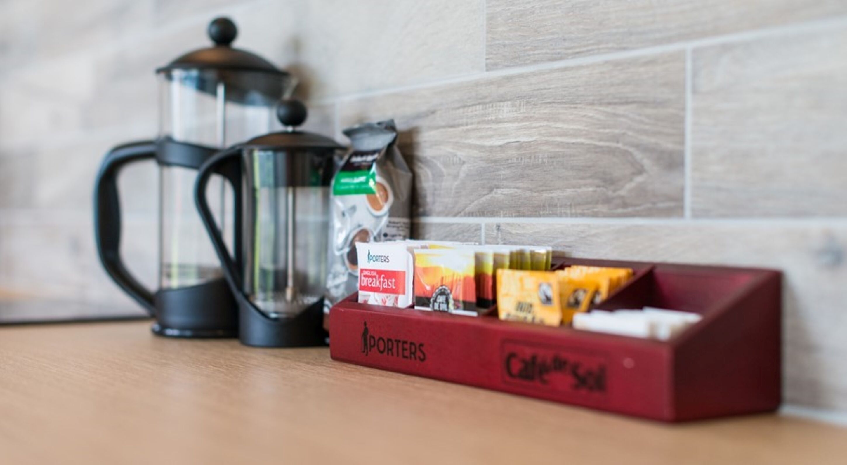 Een koffiezetapparaat, een glazen kan en een doos koffiekapsules op een houten tafel, met een betegelde muur op de achtergrond.