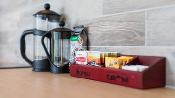 Een koffiezetapparaat, een glazen kan en een doos koffiekapsules op een houten tafel, met een betegelde muur op de achtergrond.