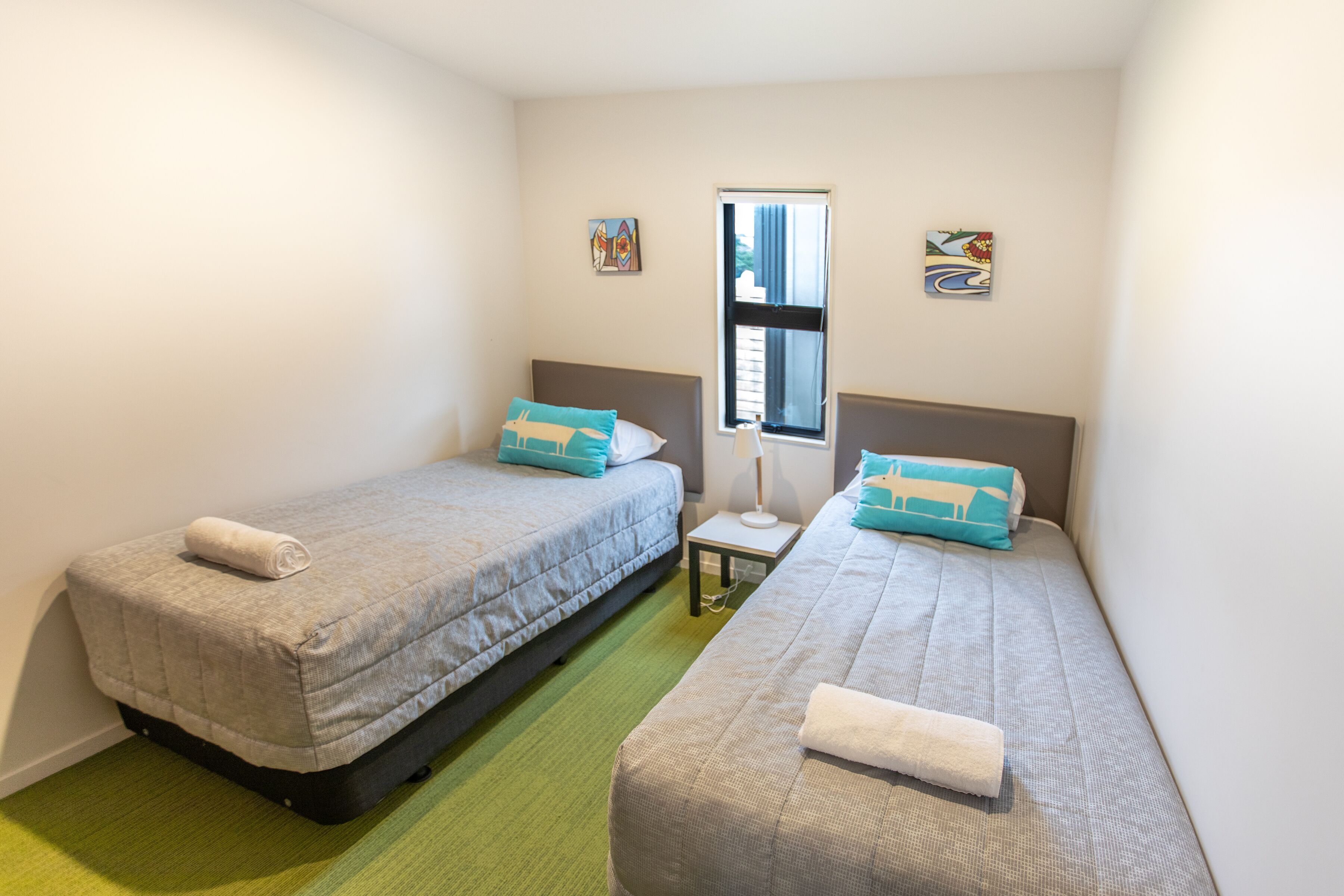 Een kleine, nette kamer met twee eenpersoonsbedden, een raam met gordijnen en een klein tafeltje met een lamp.