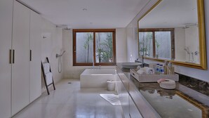 A modern bathroom with a bathtub, a large mirror, and a window with a view of trees. - Villa Nikara (Canggu)