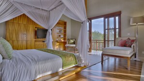 A bedroom with a canopy bed, wooden flooring, a television, a chair, and a balcony with a view of trees. - Villa Nikara (Canggu)