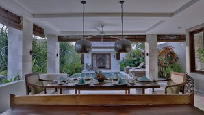 A dining area with a wooden table set for four, two hanging pendant lights, and a view of a house with a pool in the background. - Villa Nikara (Canggu)