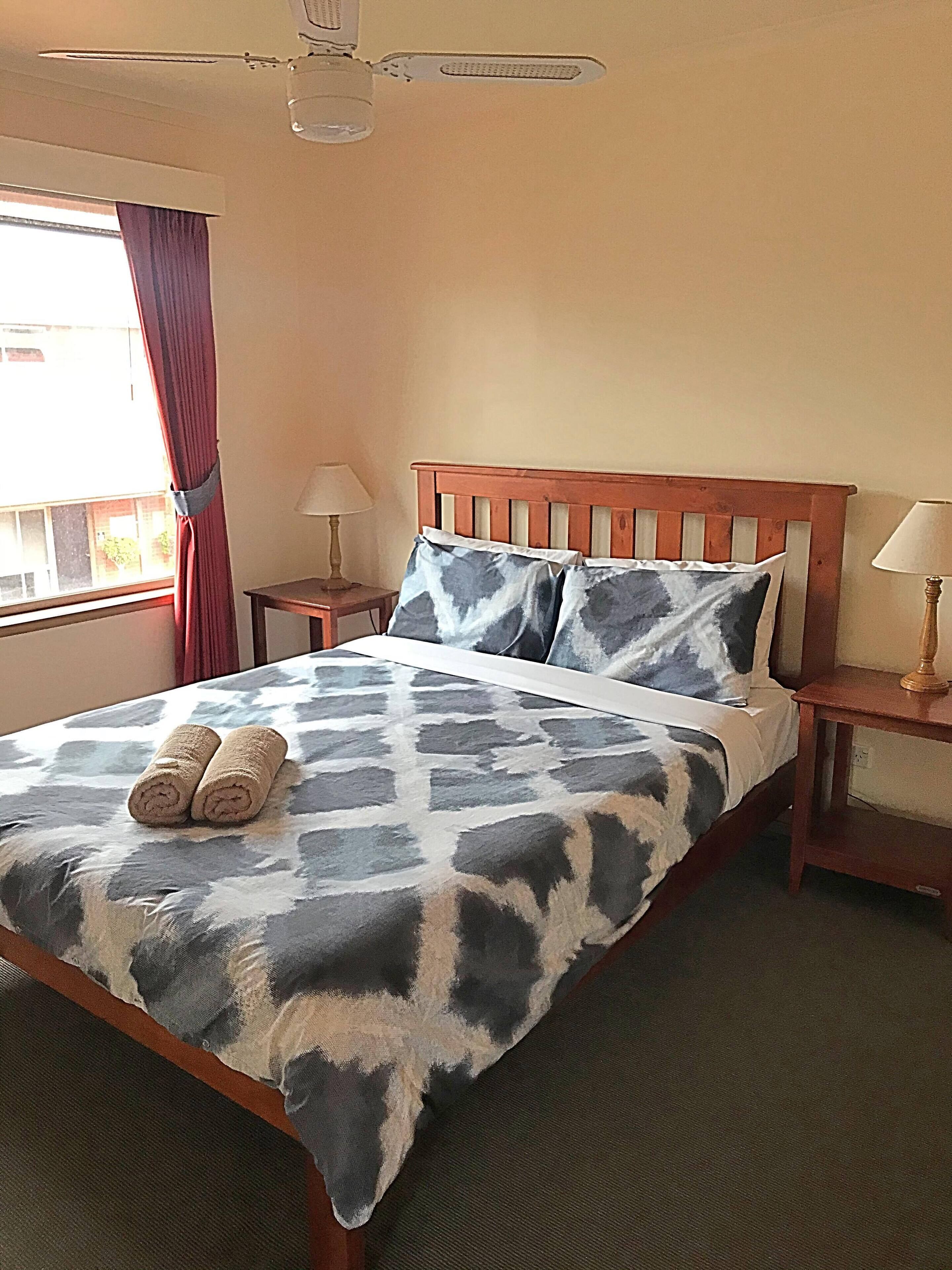 Habitación de hotel con cama, dos mesitas de noche, ventilador de techo, ventana con cortinas y vistas al exterior.