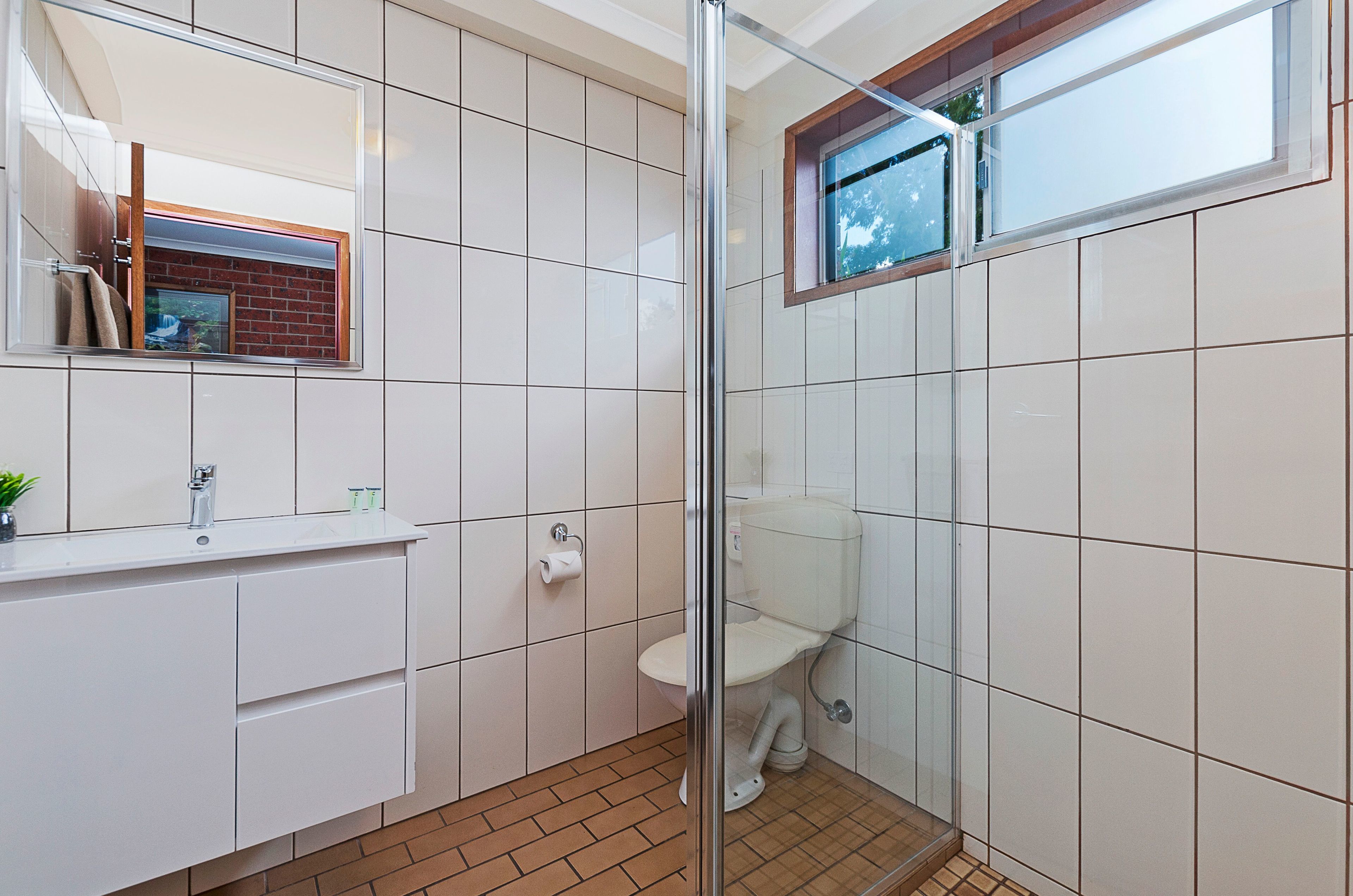 A bathroom with a glass shower enclosure, a toilet, a sink with a mirror, and a window.