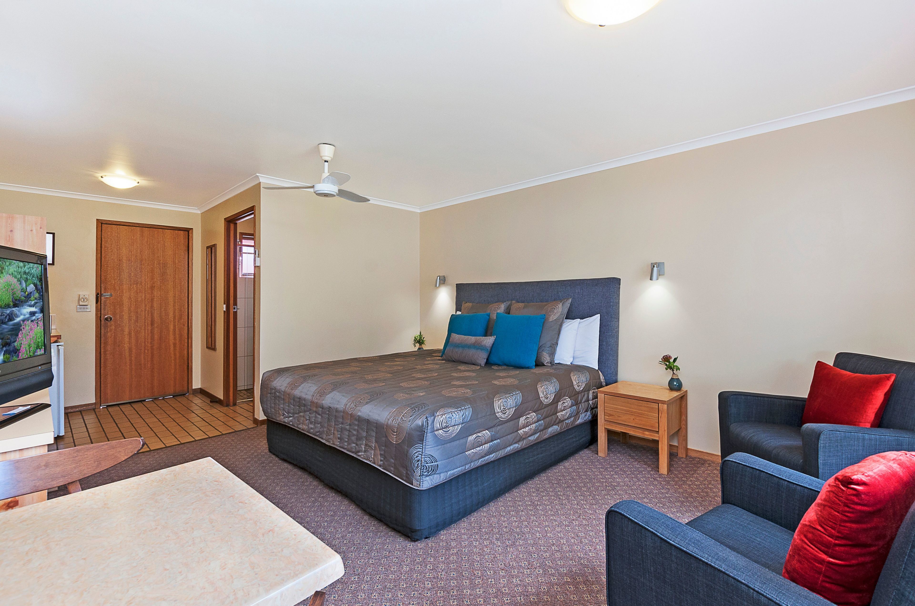 A bedroom with a bed, a TV, a ceiling fan, a nightstand, a chair, and a small table.
