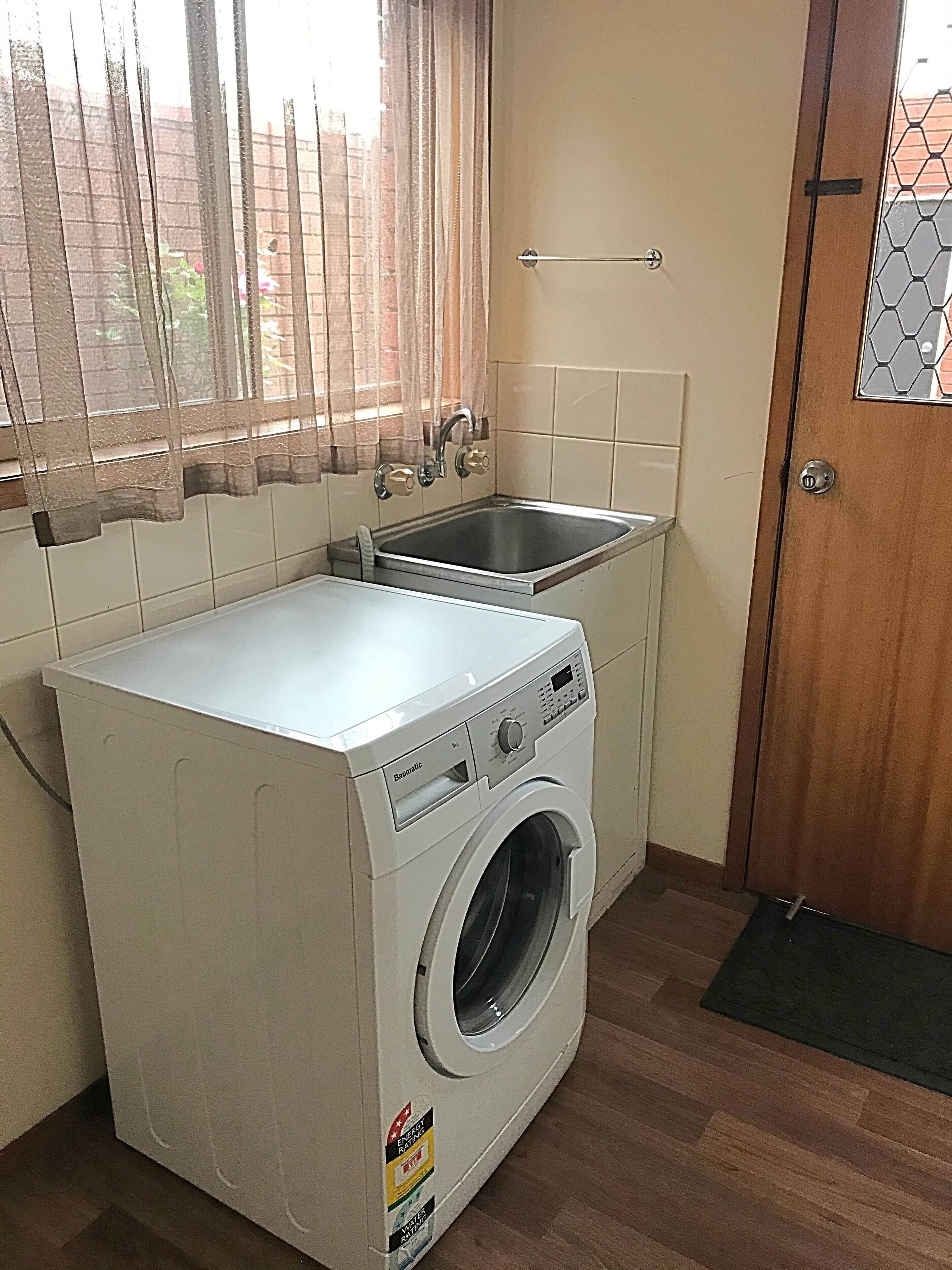 A compact laundry area with a front-loading washing machine, a sink, and a window with curtains.