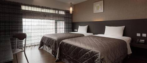 A hotel room with two beds, a chair, a desk, and a large window with patterned curtains.
