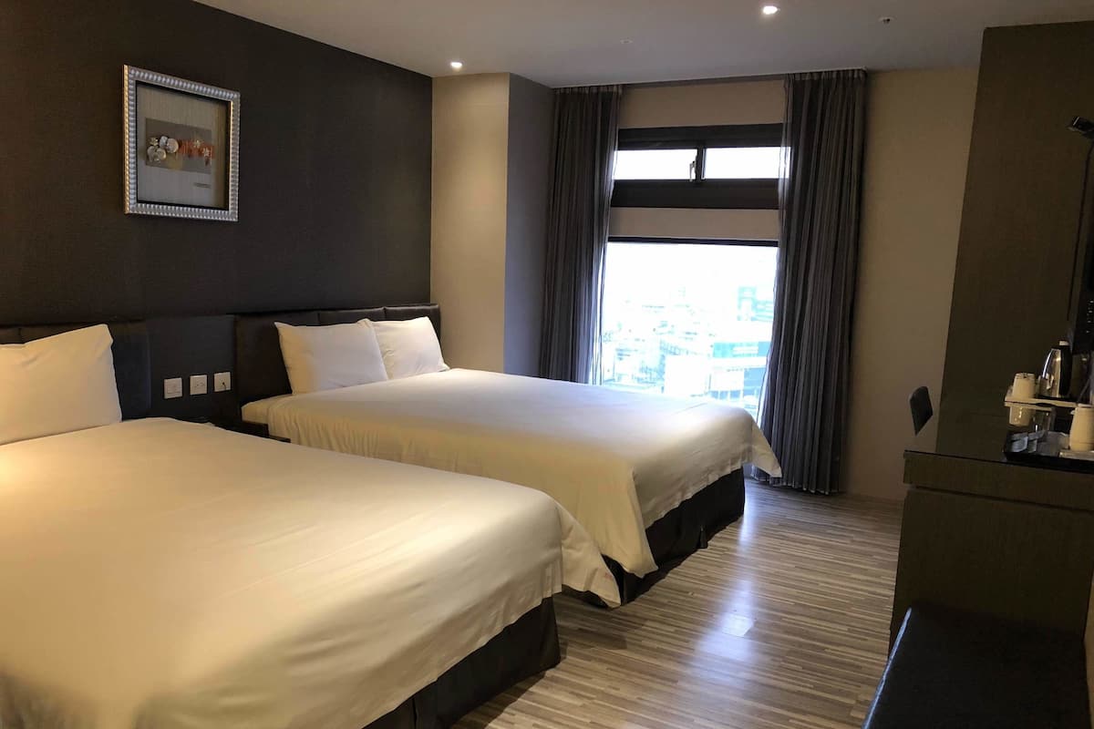 A hotel room with two beds, a large window with curtains, a framed picture on the wall, and a desk with a coffee maker.