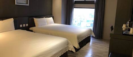 A hotel room with two beds, a large window with curtains, a framed picture on the wall, and a desk with a coffee maker.