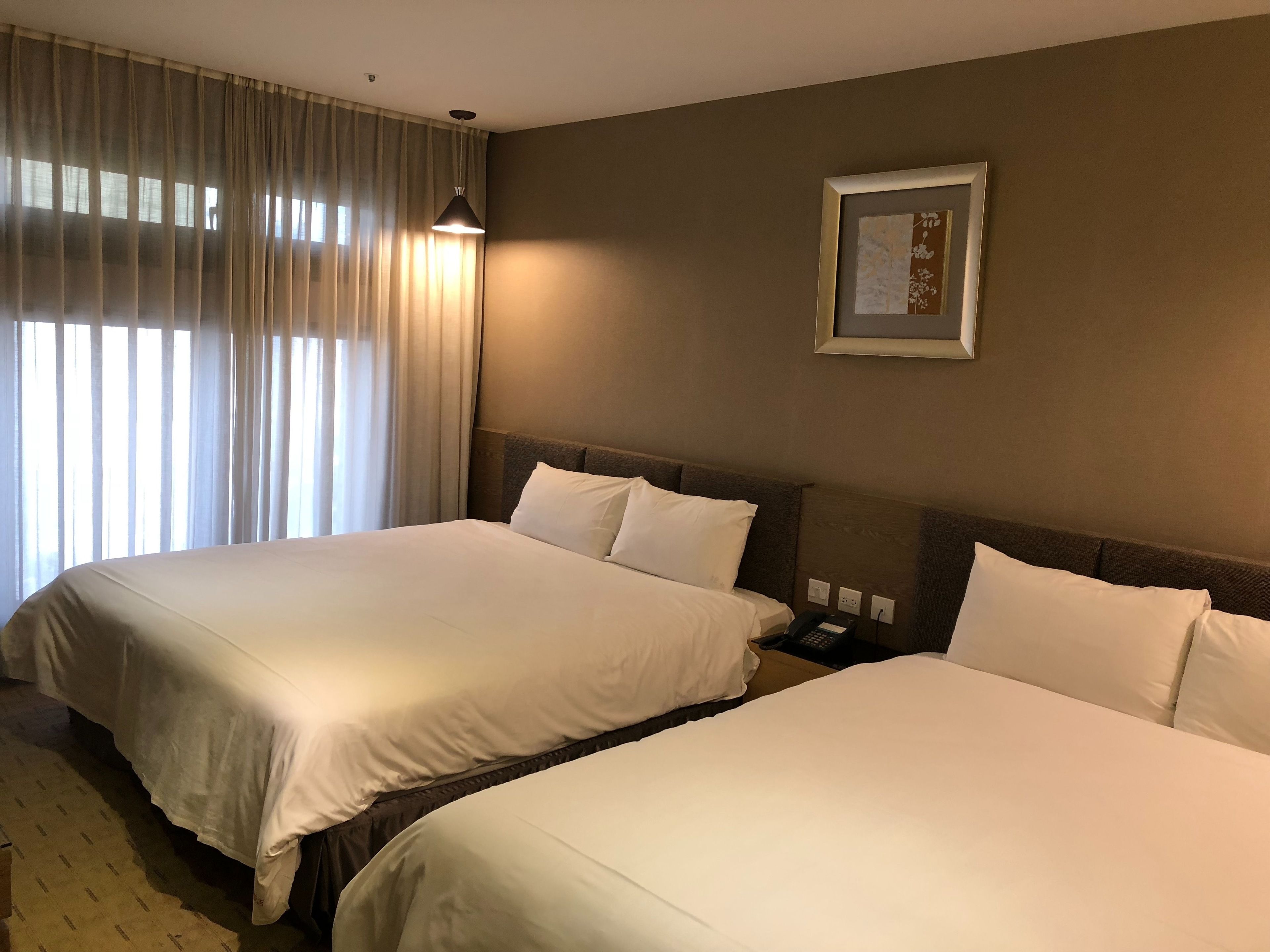 A hotel room with two beds, a large window with curtains, a wall-mounted phone, and a framed picture.