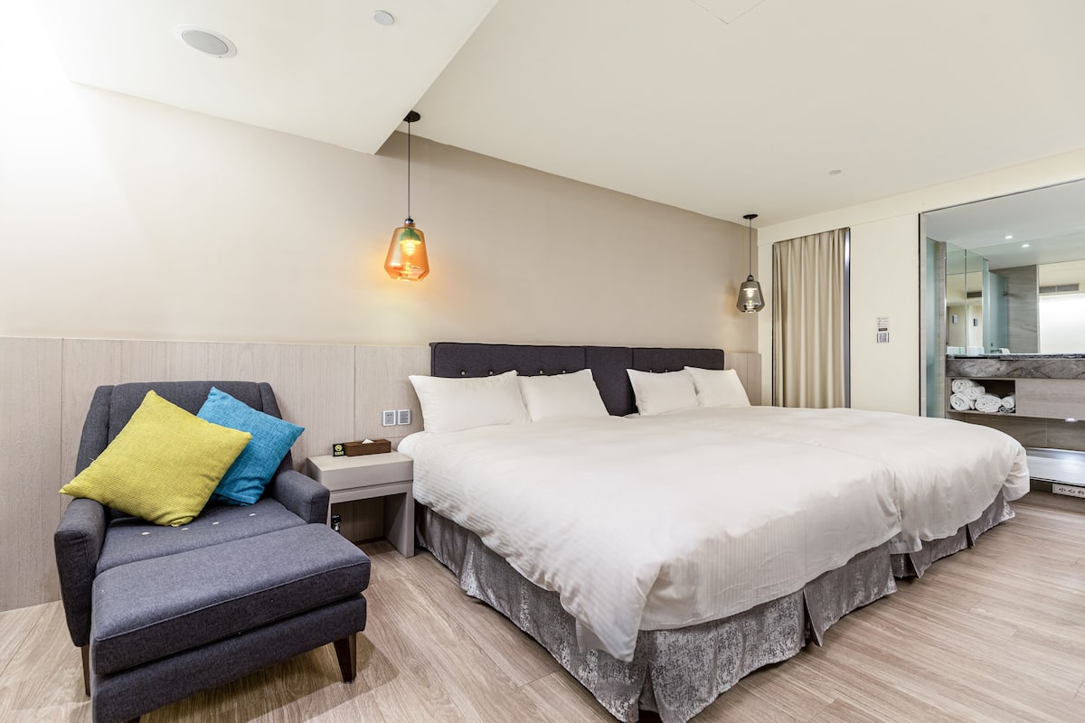 Une chambre d’hôtel moderne avec un grand lit, un fauteuil gris agrémenté de coussins, une table de chevet et un luminaire suspendu.