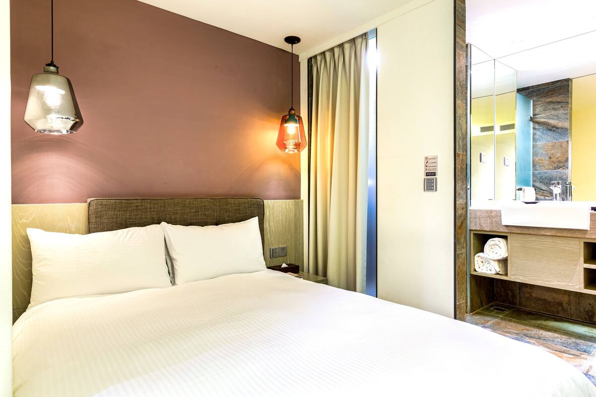 Une chambre d’hôtel avec un grand lit, deux lampes de chevet, une table de chevet, un miroir et une salle de bain équipée d’un lavabo et d’un porte-serviettes.
