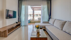 A modern living room with a sofa, a coffee table with a vase, a TV on the wall, and a view of an outdoor pool area. - The Pelican Residence & Suite Krabi (Krabi)