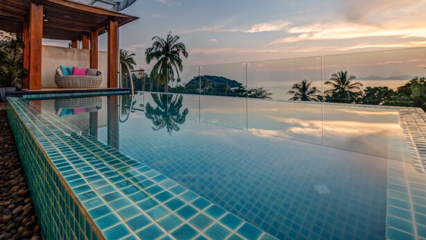 A infinity pool with a view of palm trees and a distant island, a covered seating area with cushions, and a tiled edge. - The Pelican Residence & Suite Krabi (Krabi)