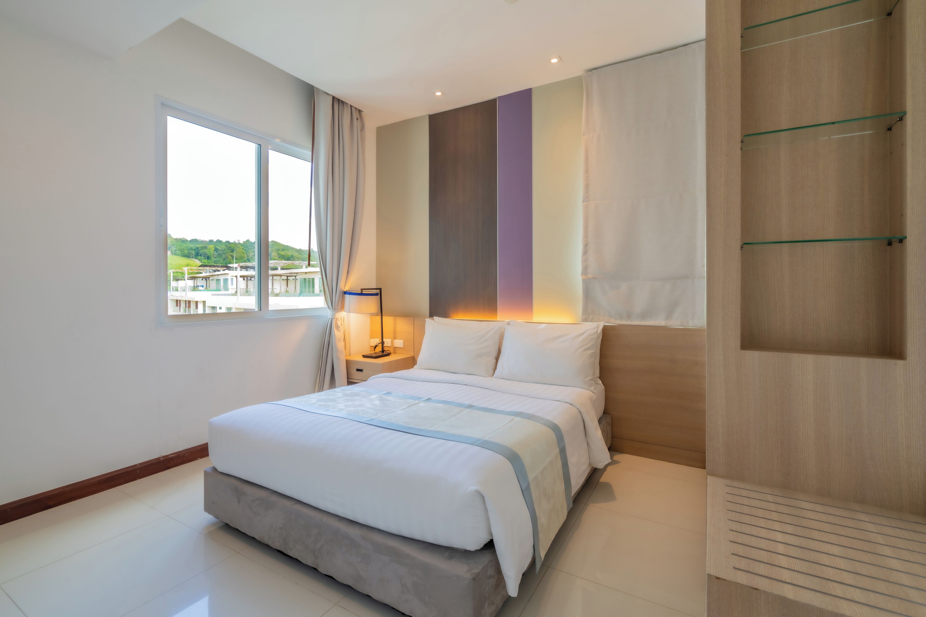 A modern bedroom with a large bed, a nightstand, a window with curtains, and a view of greenery outside.