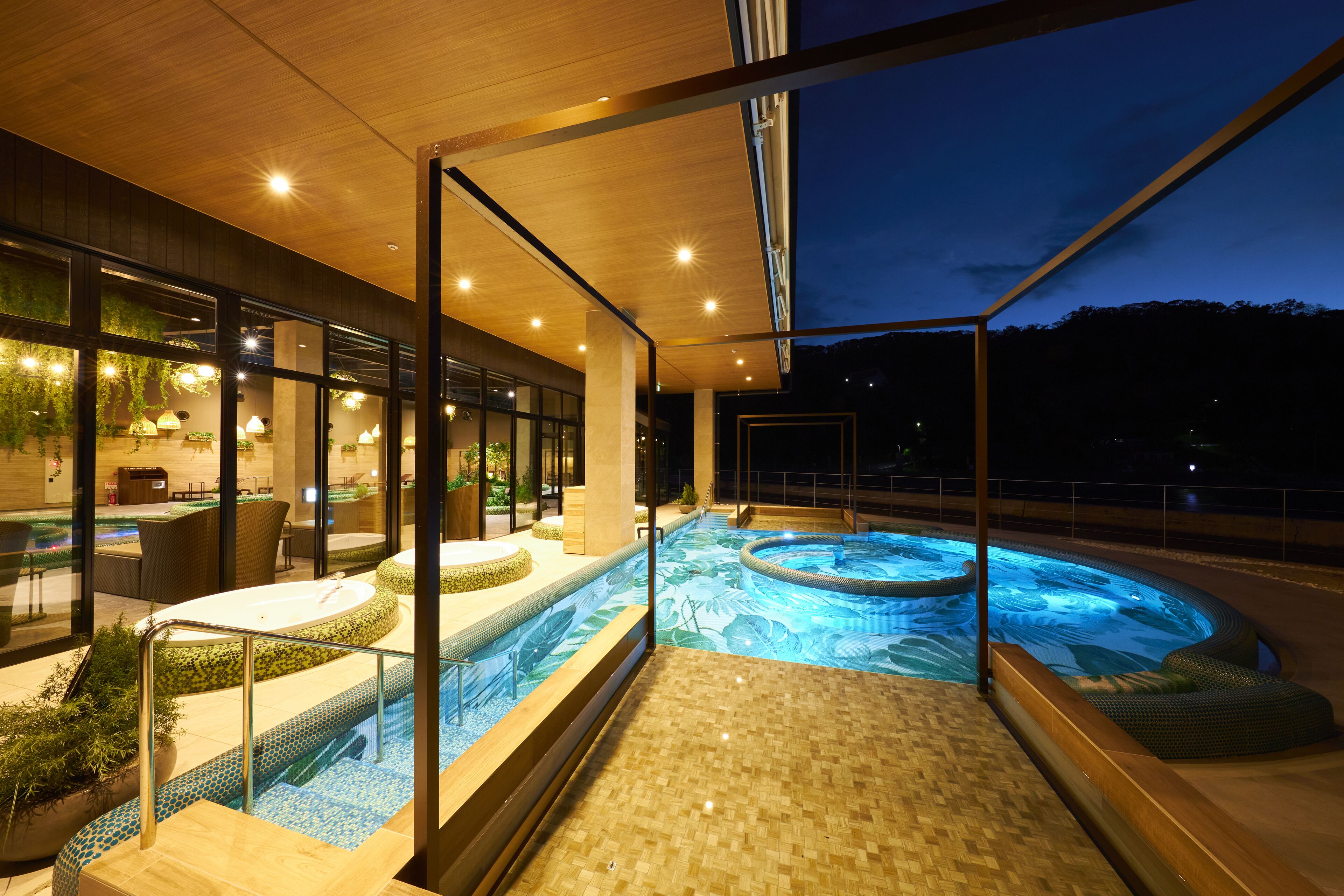 Un área de piscina interior moderna con un jacuzzi, asientos y vistas al cielo nocturno.