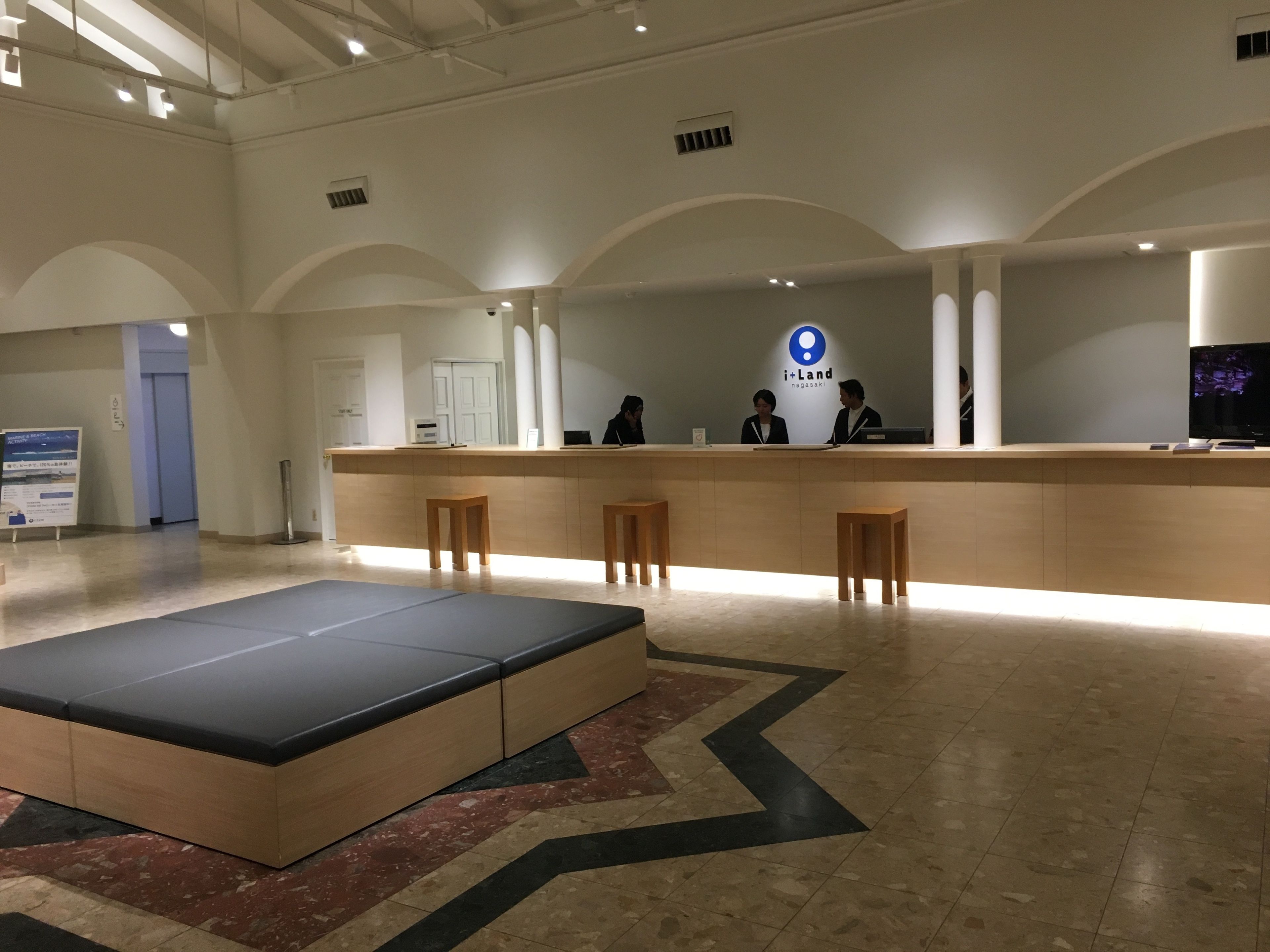 Un área de recepción con mostrador, tres taburetes de madera, un sofá grande negro y beige, y un piso estampado.