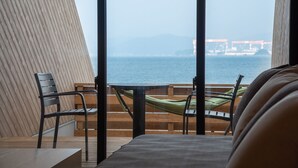 A room with a large window overlooking the sea, a bed, a table, and two chairs.