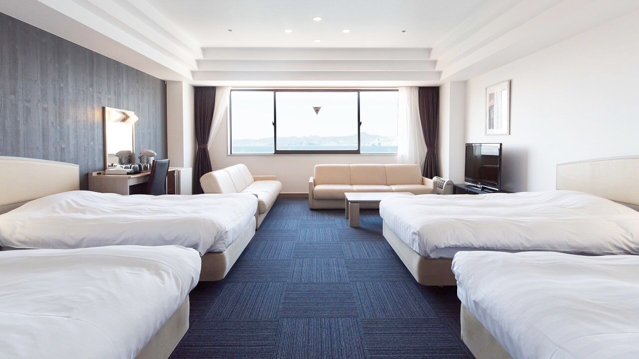 A hotel room with two beds, a sofa, a desk, a TV, and a large window.