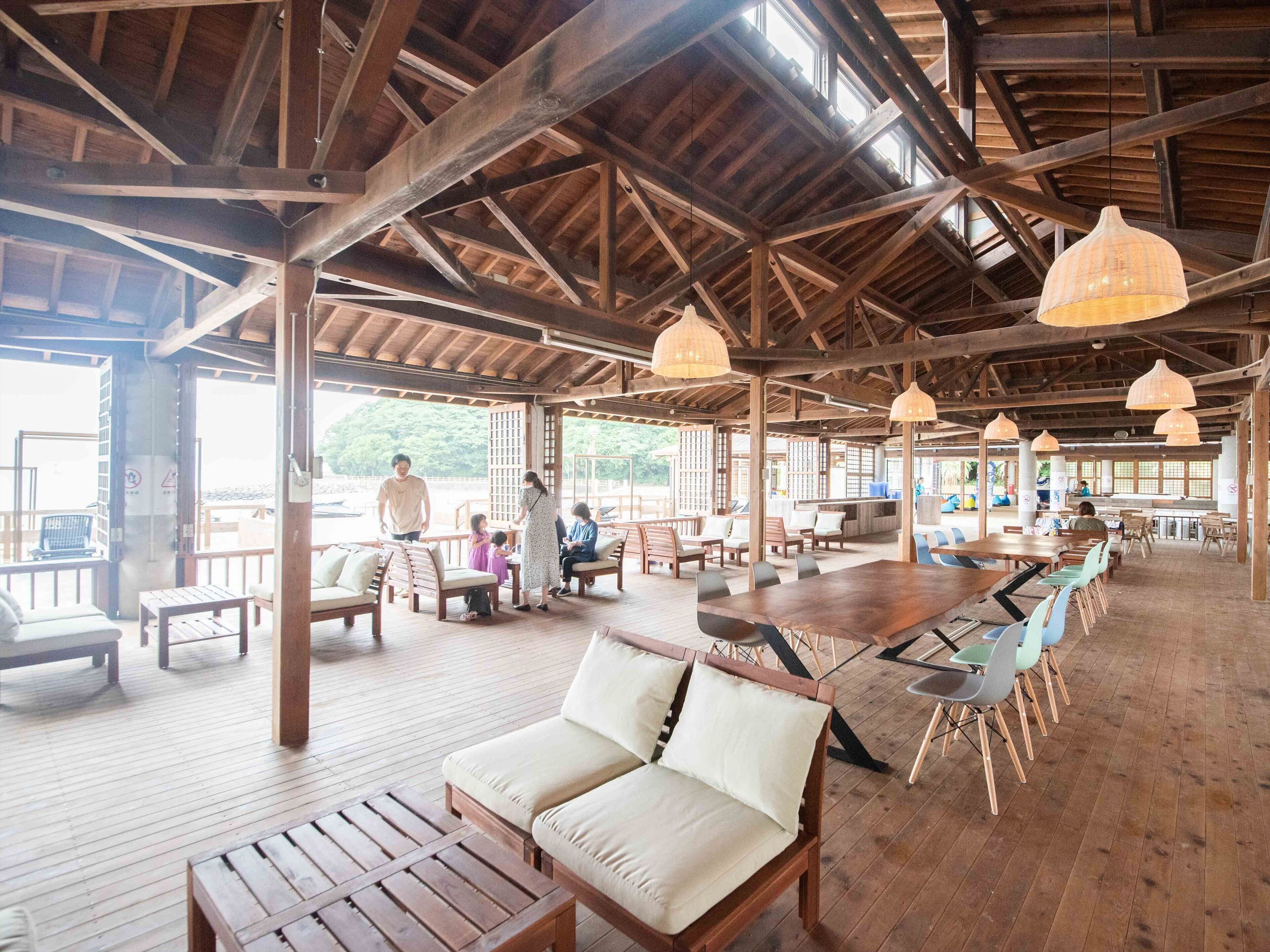 A spacious outdoor dining area with wooden tables and chairs, a covered seating area with cushioned benches, and hanging pendant lights.