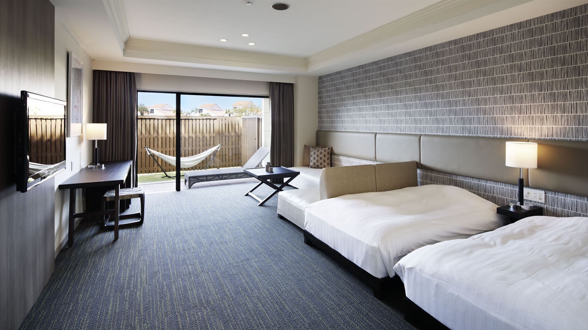 A hotel room with two beds, a desk, a chair, a hammock, and a view of the outside.