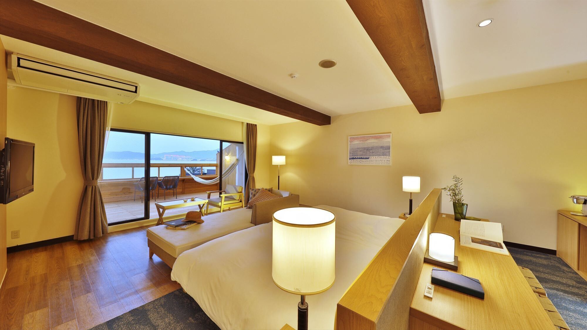 A hotel room with a bed, bedside table, desk, chair, and a large window offering a view.