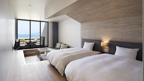 A modern hotel room with two beds, a large window with a view of the sea, a seating area with a table, and a wooden accent wall.