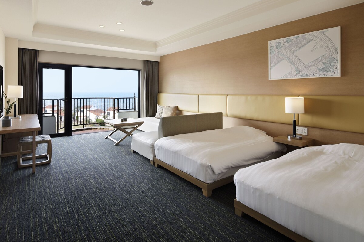 A hotel room with two beds, a desk, a chair, a small table, a lamp, and a large window with a view of the sea.
