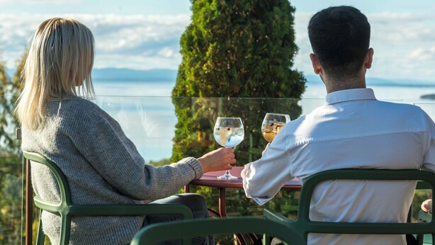 Twee personen zitten aan een tafel met een glas wijn, met uitzicht op een schilderachtig landschap met een hoge boom en een wateroppervlak.