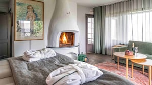 Een moderne hotelkamer met een open haard, een bed met een grijze deken, een groene tas en een klein, rond tafeltje met een fles en glazen.