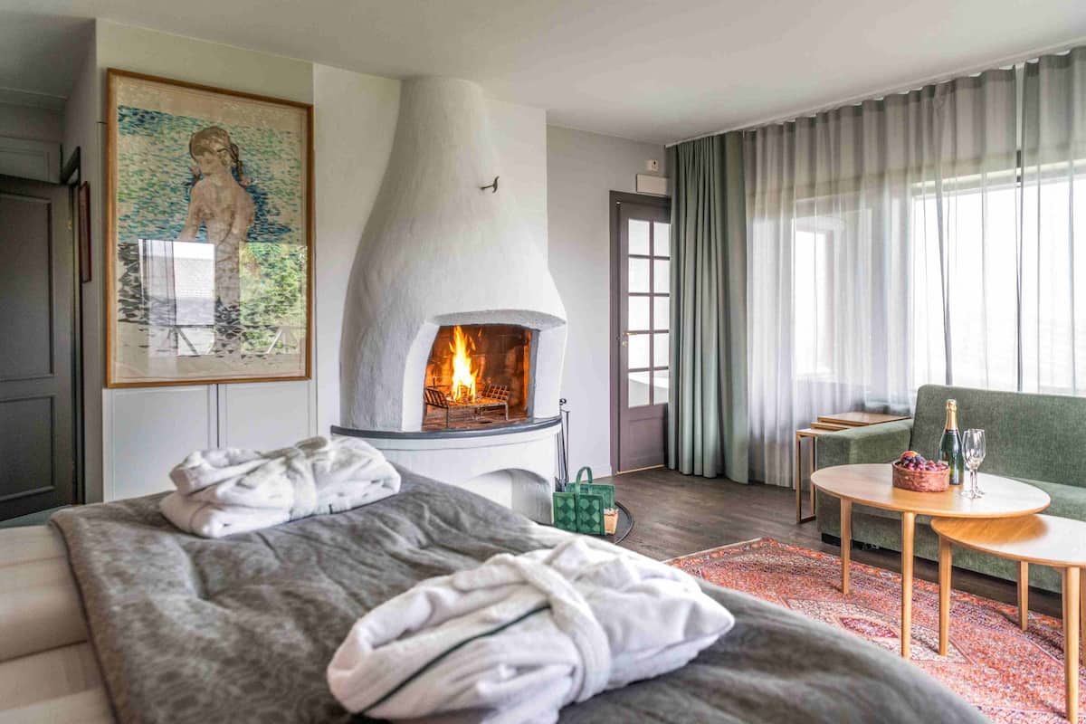 Una habitación de hotel moderna con chimenea, una cama con manta gris, un bolso verde y una pequeña mesa redonda con una botella y vasos.