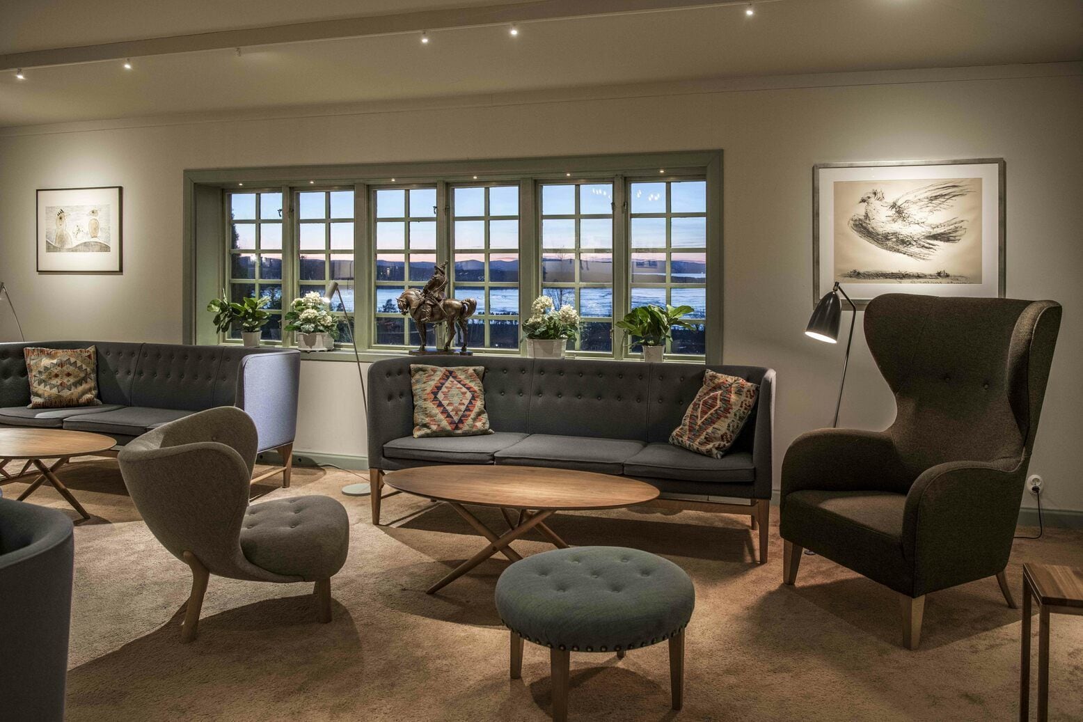 Una sala de estar moderna con un sofá, sillones, una mesa de centro redonda y vistas al exterior a través de grandes ventanales.