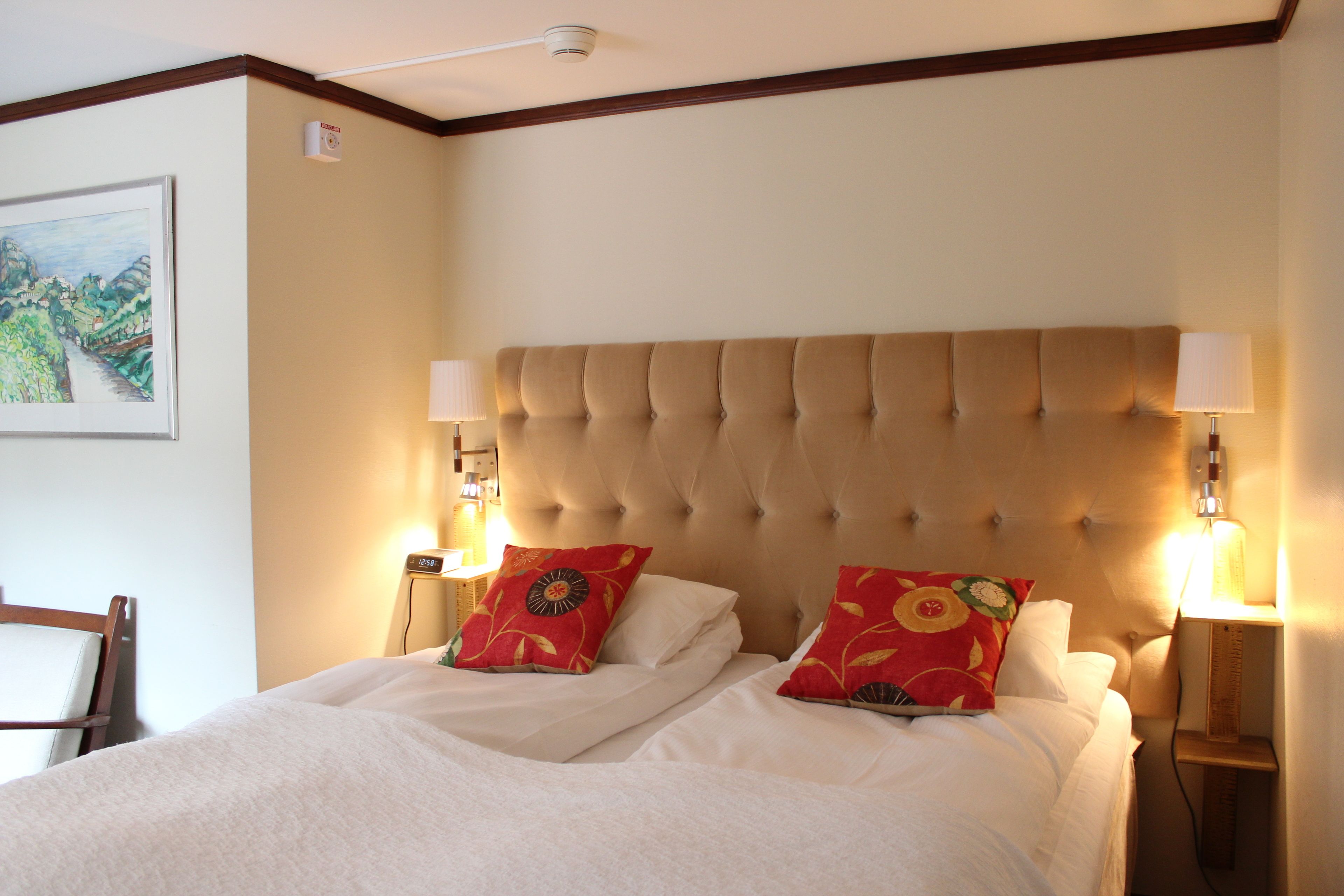 Una cama bien hecha con un cabecero capitonado, dos lámparas de noche y dos almohadas rojas con diseños florales.